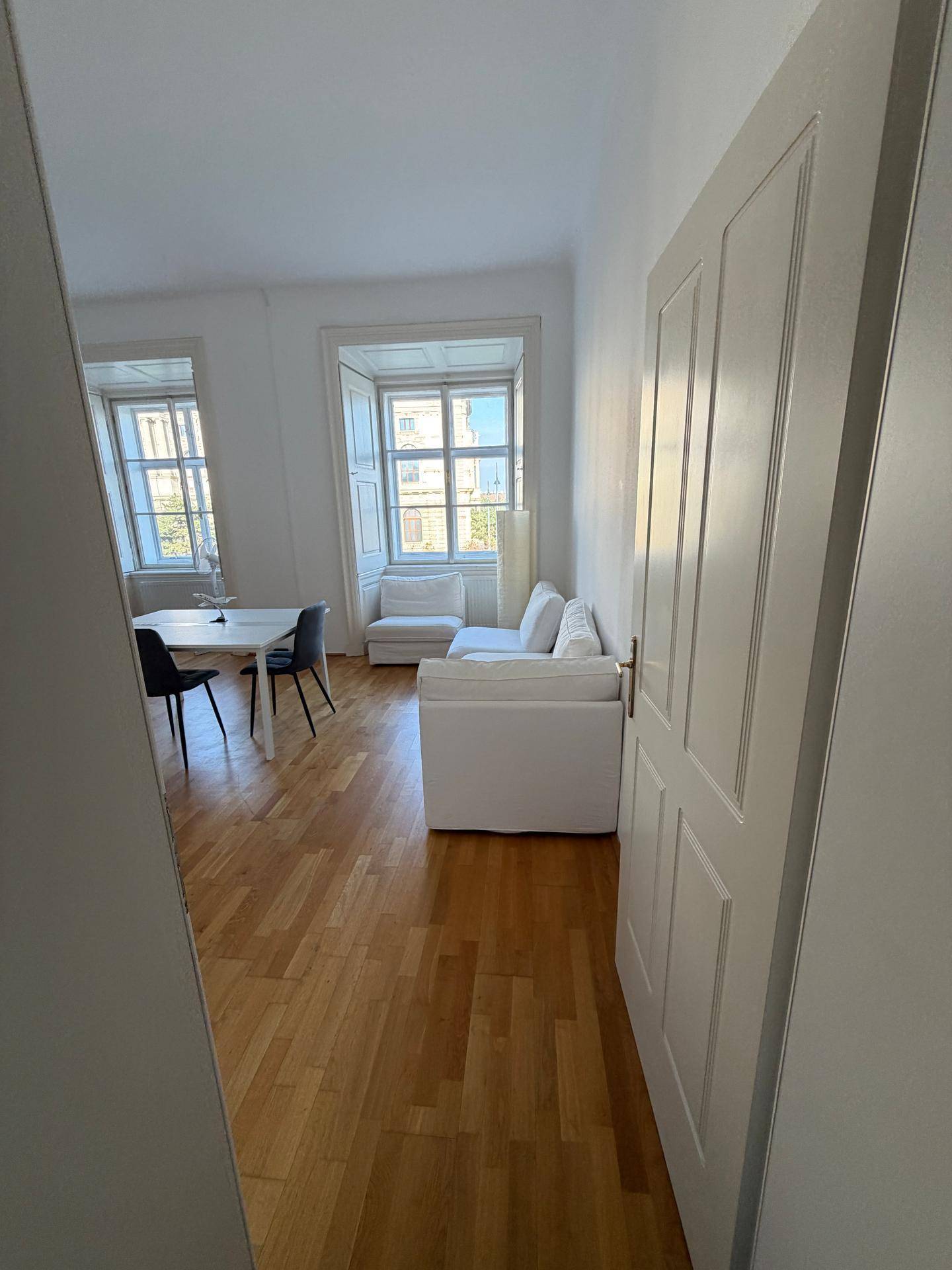 Geräumiges Wohnzimmer mit hellen Wänden, Holzfußboden und großen Fenstern, vom Flur aus sichtbar.