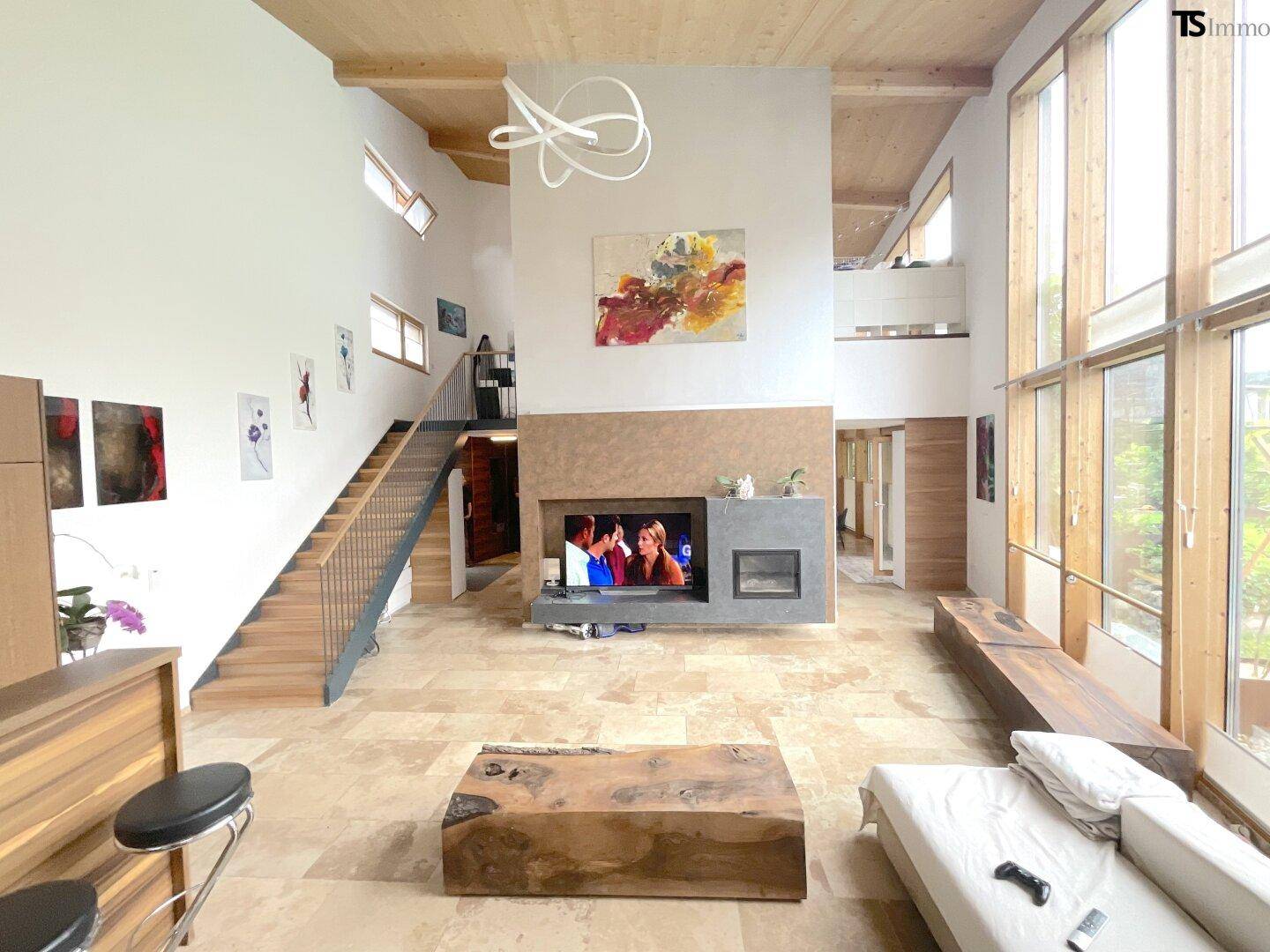 Großzügiges Wohnzimmer mit hohen Decken, Kamin, Steinboden und großen Fenstern zum Garten.