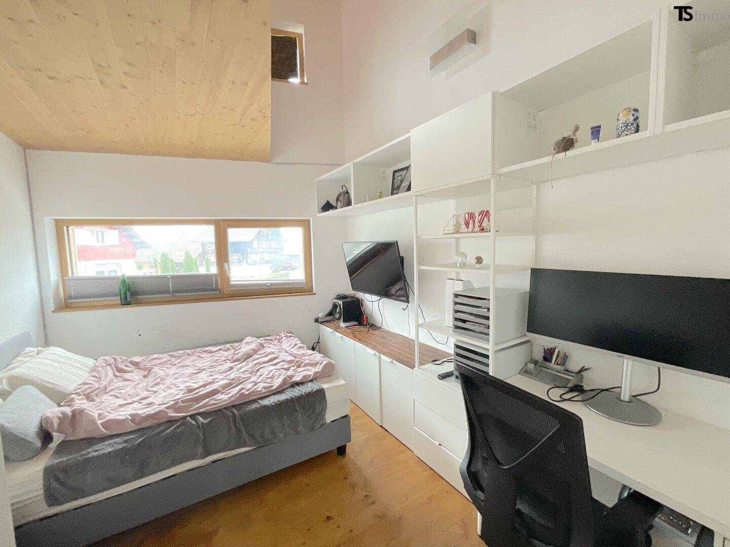 Helles Schlafzimmer mit Arbeitsbereich, Holzdecke und Blick ins Grüne durch das Fenster.