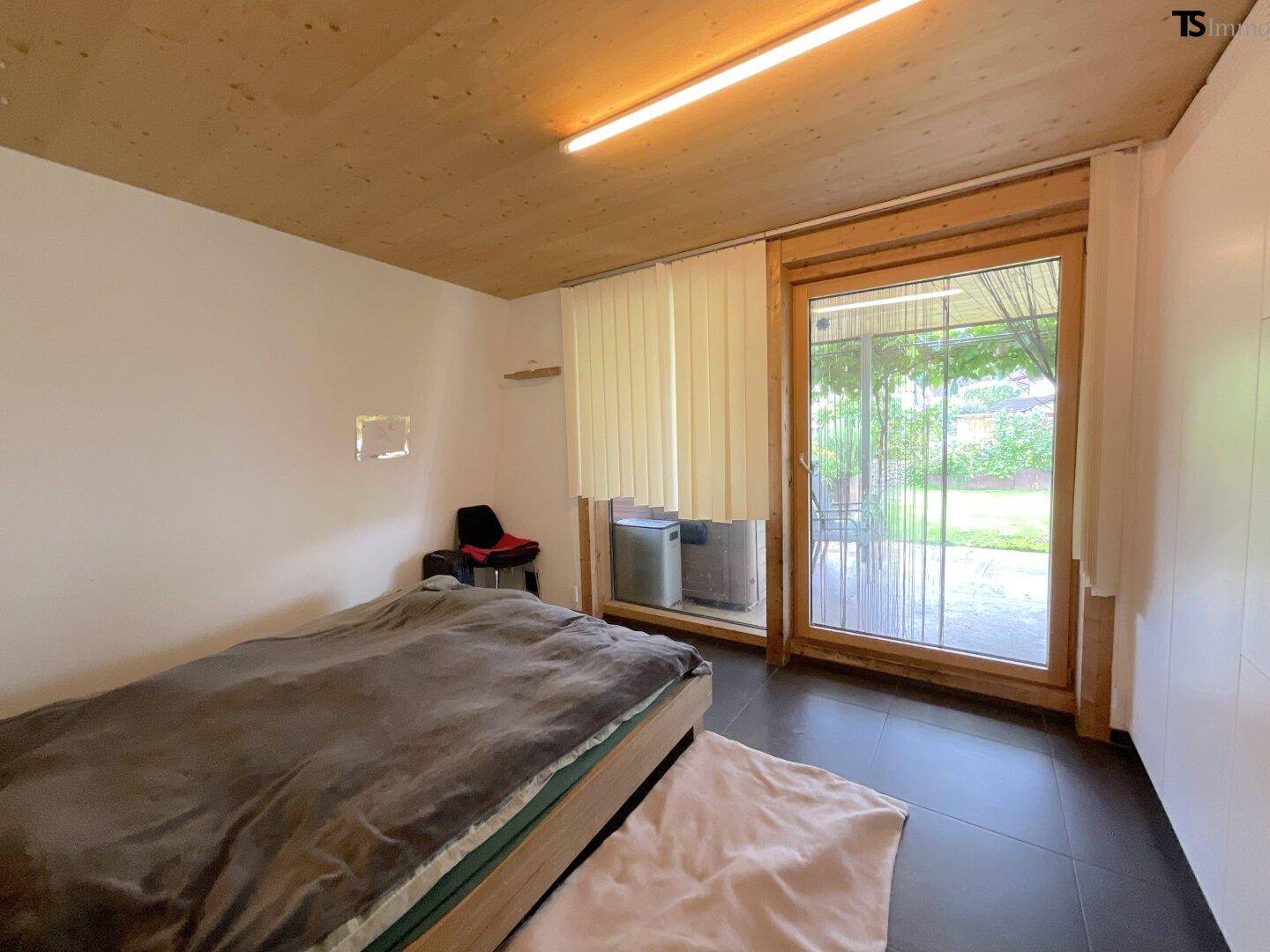 Schlafzimmer mit direktem Zugang zum Garten, dunklem Fliesenboden und großen Fenstern.