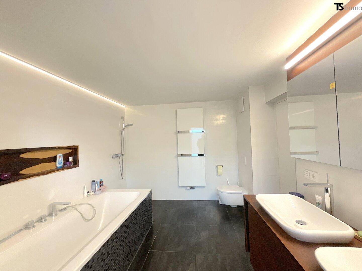 Modernes Badezimmer mit Badewanne, Einzelwaschbecken, dunklen Fliesen und indirekter Beleuchtung.