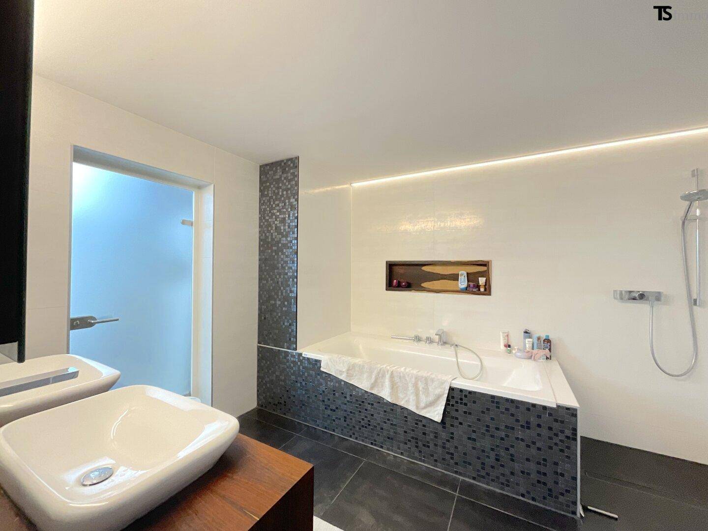Geräumiges modernes Badezimmer mit Badewanne, Doppelwaschbecken und dunklen Mosaikfliesen.