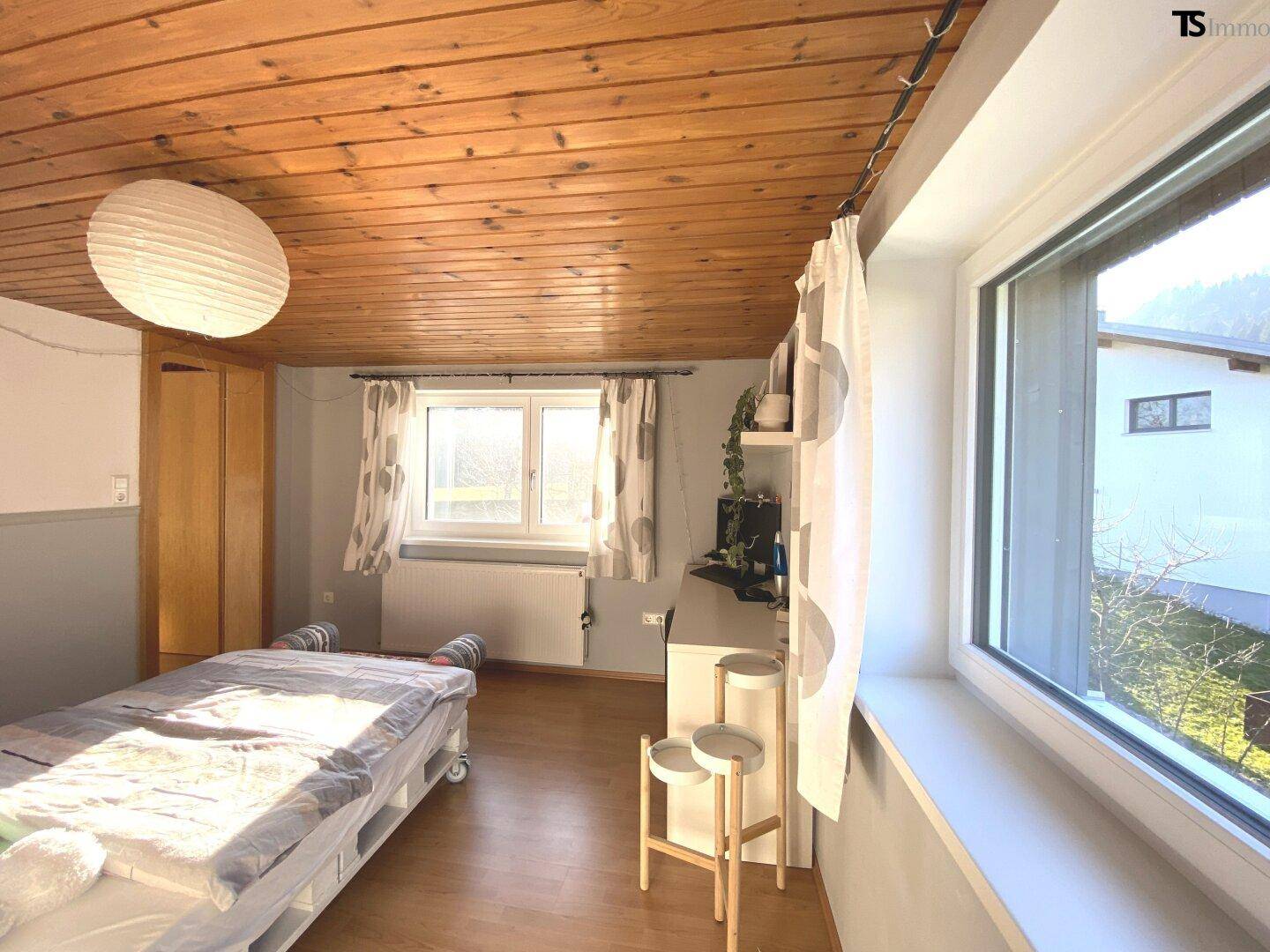 Gemütliches Schlafzimmer mit Holzdecke, Holzboden und viel Tageslicht durch die Fenster.