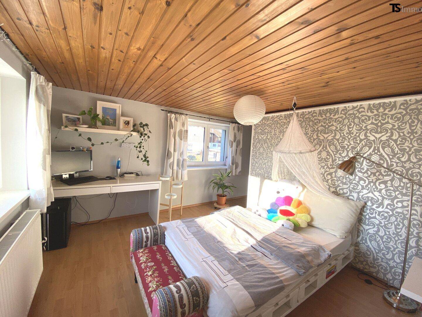 Helles Schlafzimmer mit Holzdecke, Schreibtisch und dekorativer Tapete an der Wand.