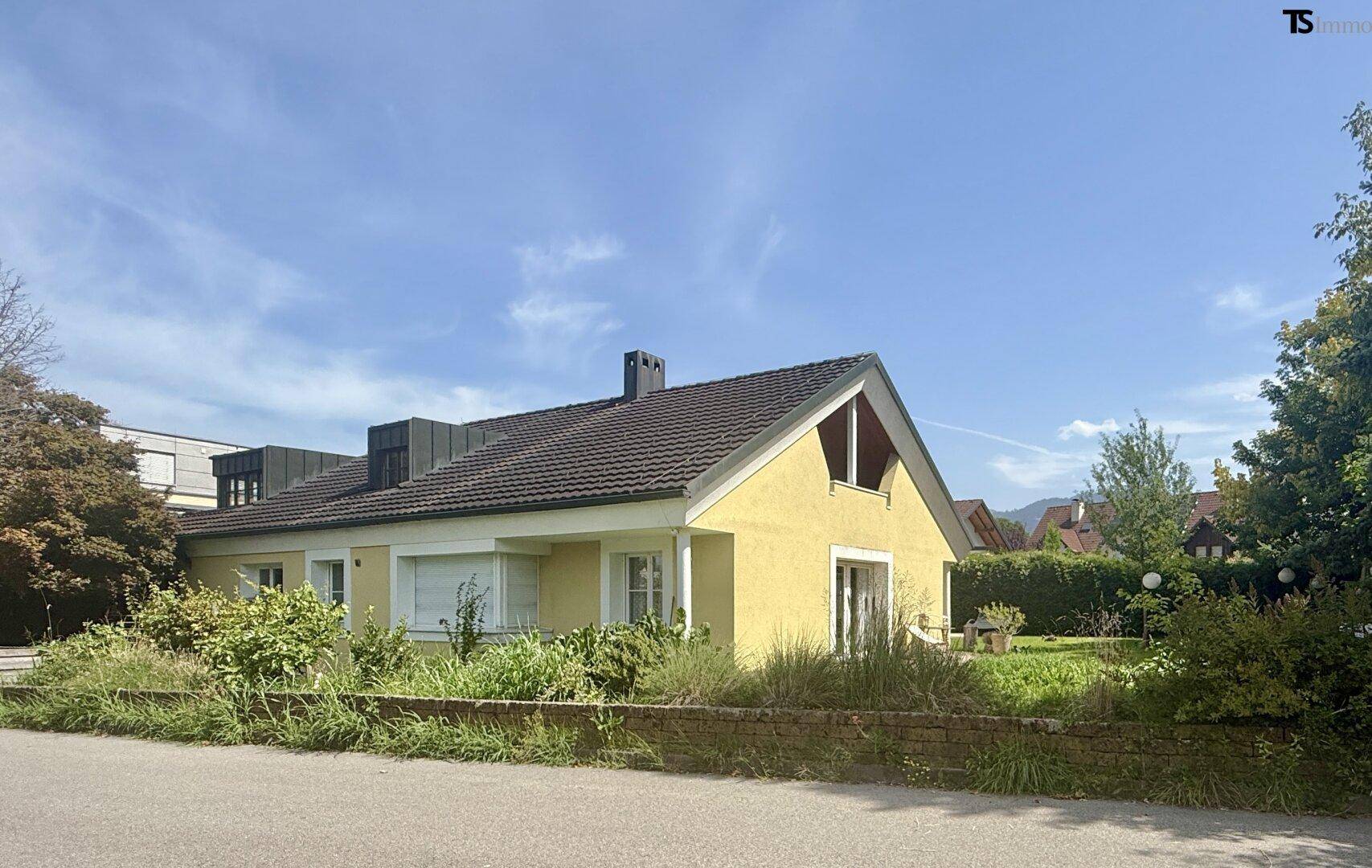 Gelbes Einfamilienhaus mit Satteldach und Vorgarten unter blauem Himmel.