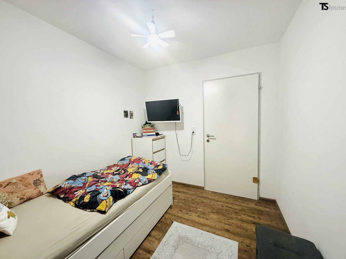 Praktisches Schlafzimmer mit wandmontiertem Fernseher und hellem Holzfußboden.