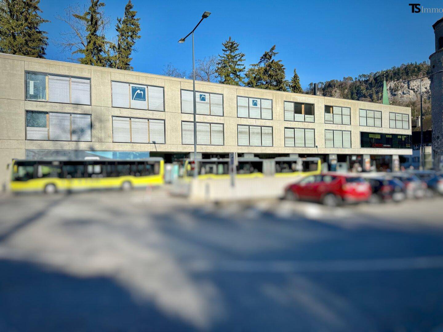 Modernes Bürogebäude mit großen Fenstern und Bussen davor, umgeben von Bäumen und einem Berg.