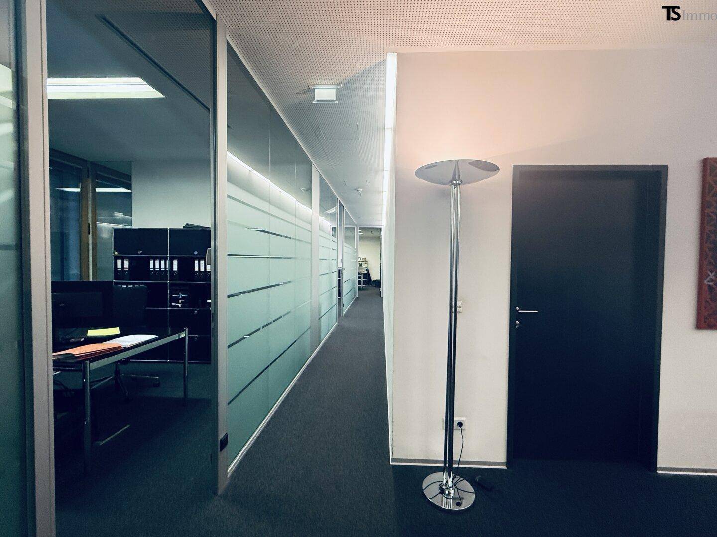 Langer Bürogang mit Glaswänden und undurchsichtigen Streifen, der zu weiteren Büroräumen führt.
