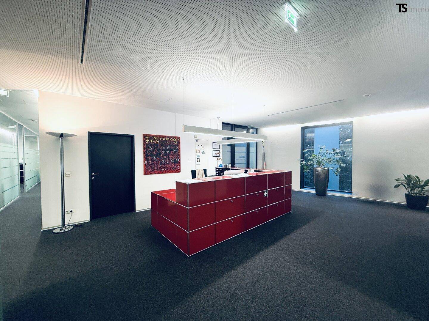 Moderner Empfangsbereich in einem Bürogebäude mit rotem Möbelstück und großen Fenstern, die Tageslicht hereinlassen.