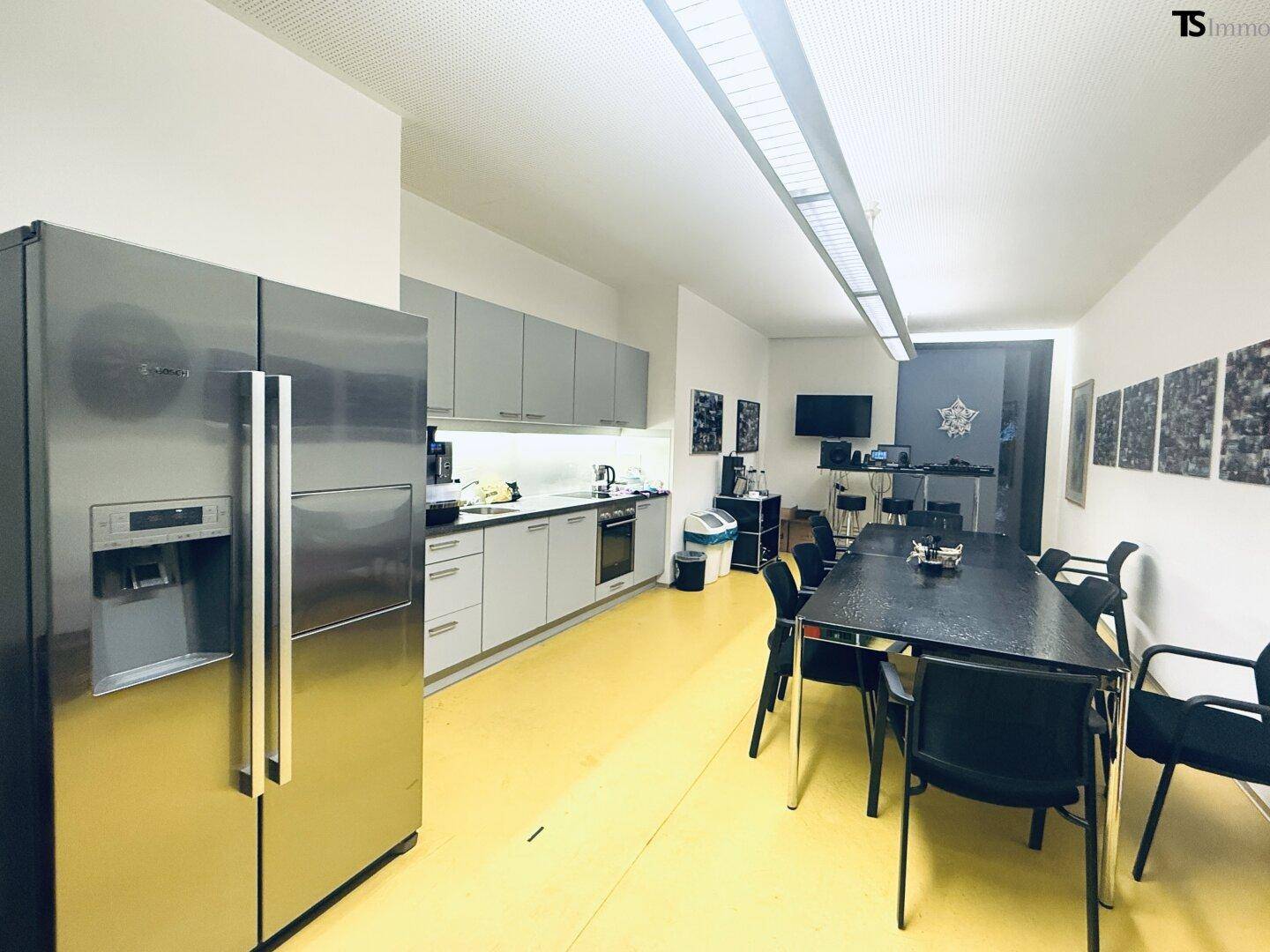Moderne Büroküche mit Edelstahlkühlschrank, Esstisch und gelbem Boden, ideal für Pausen.