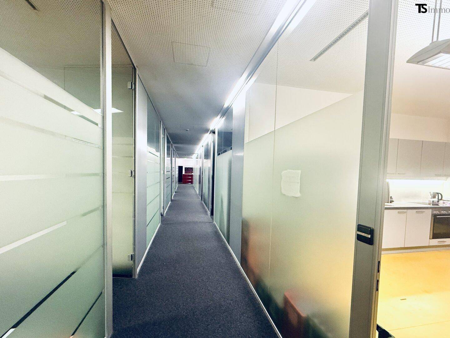 Langer Bürogang mit Glaswänden, der zu einer modernen Küche mit gelbem Boden führt.