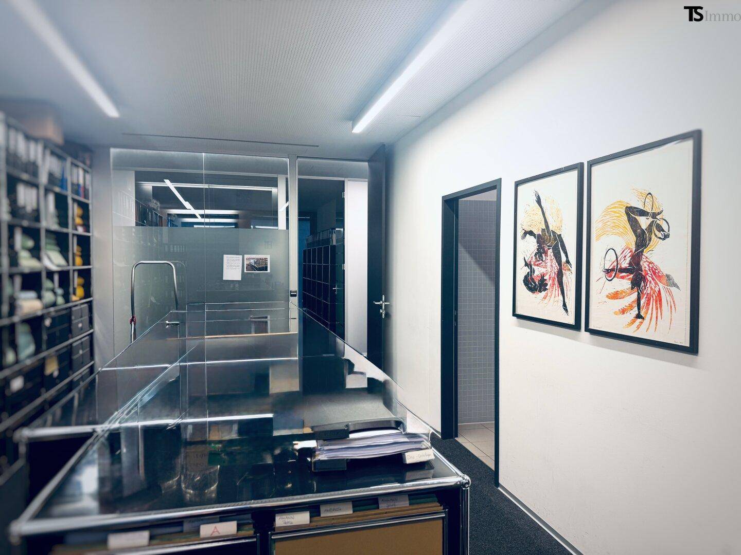 Büroflur mit Kunstwerken an der Wand und Blick in einen angrenzenden Raum mit Regalen.