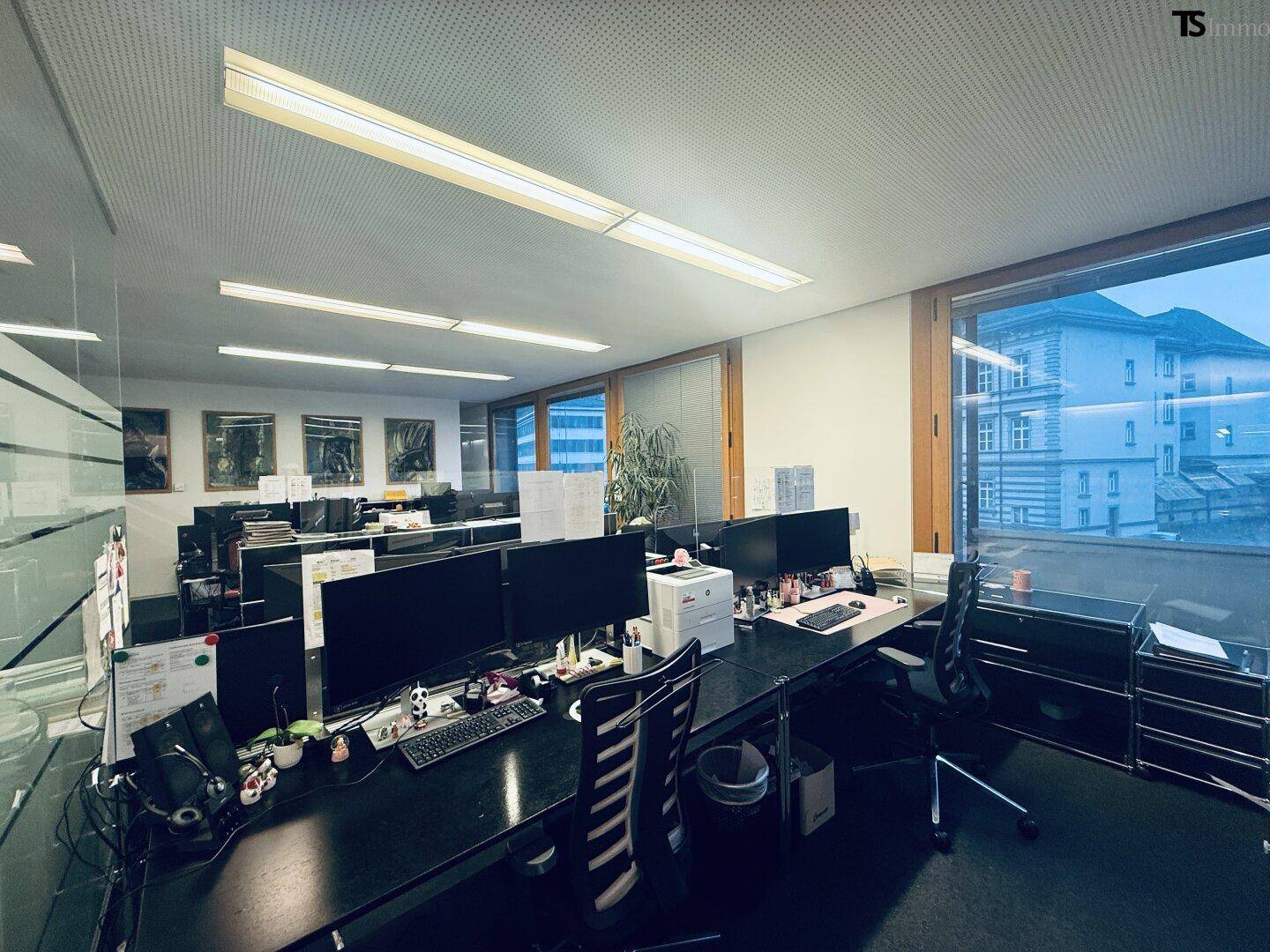 Großraumbüro mit mehreren Arbeitsplätzen, Computermonitoren und Blick auf die umliegenden Gebäude.