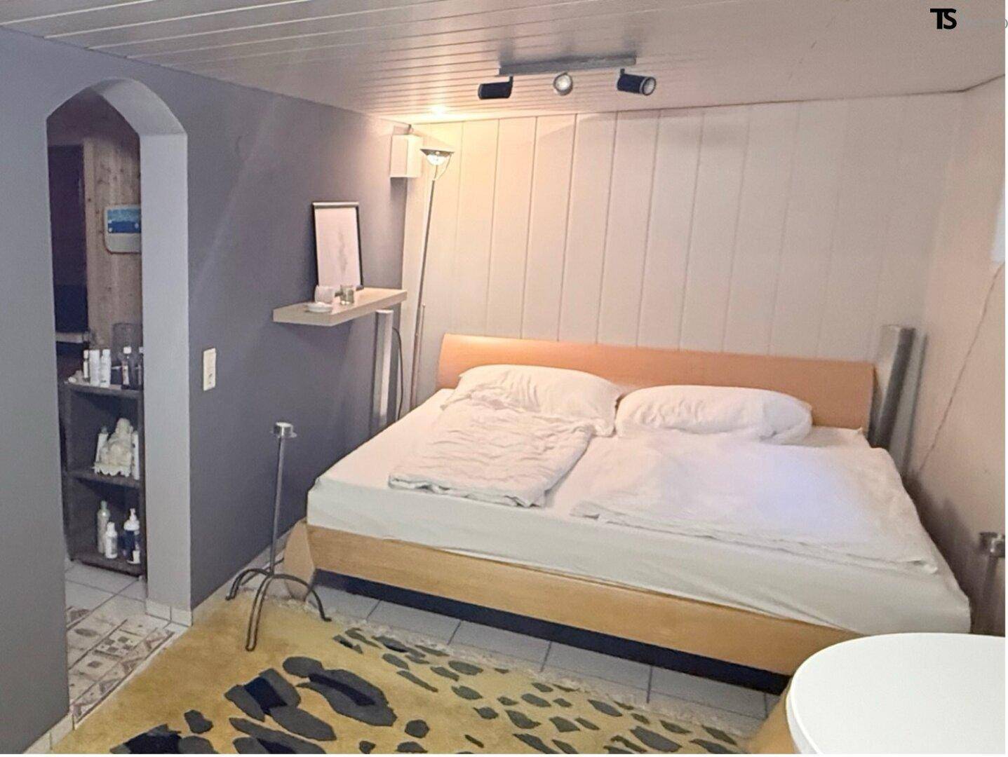 Schlafzimmer mit Holzbett, Teppich und Zugang zu einem weiteren Bereich.
