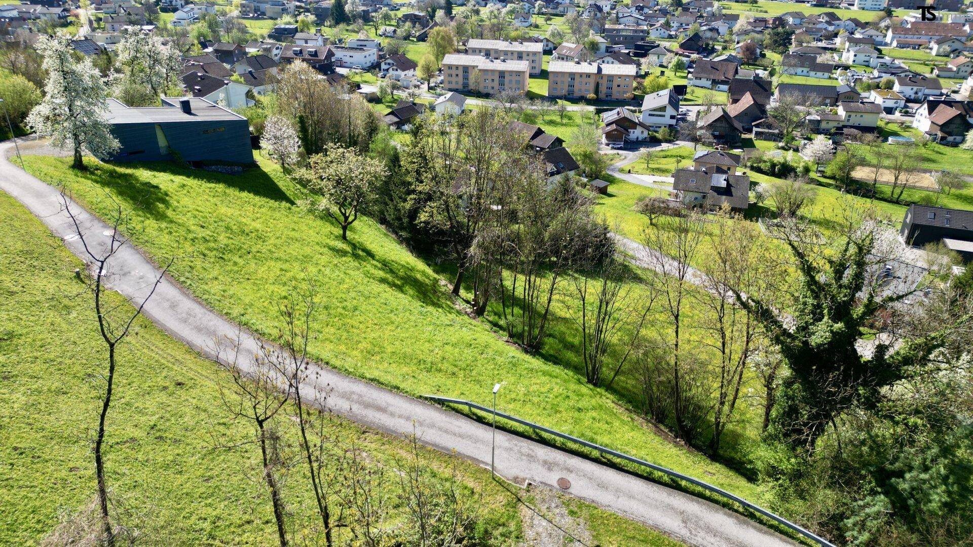 Luftaufnahme eines weitläufigen grünen Geländes mit einem Weg und Blick auf eine Siedlung.