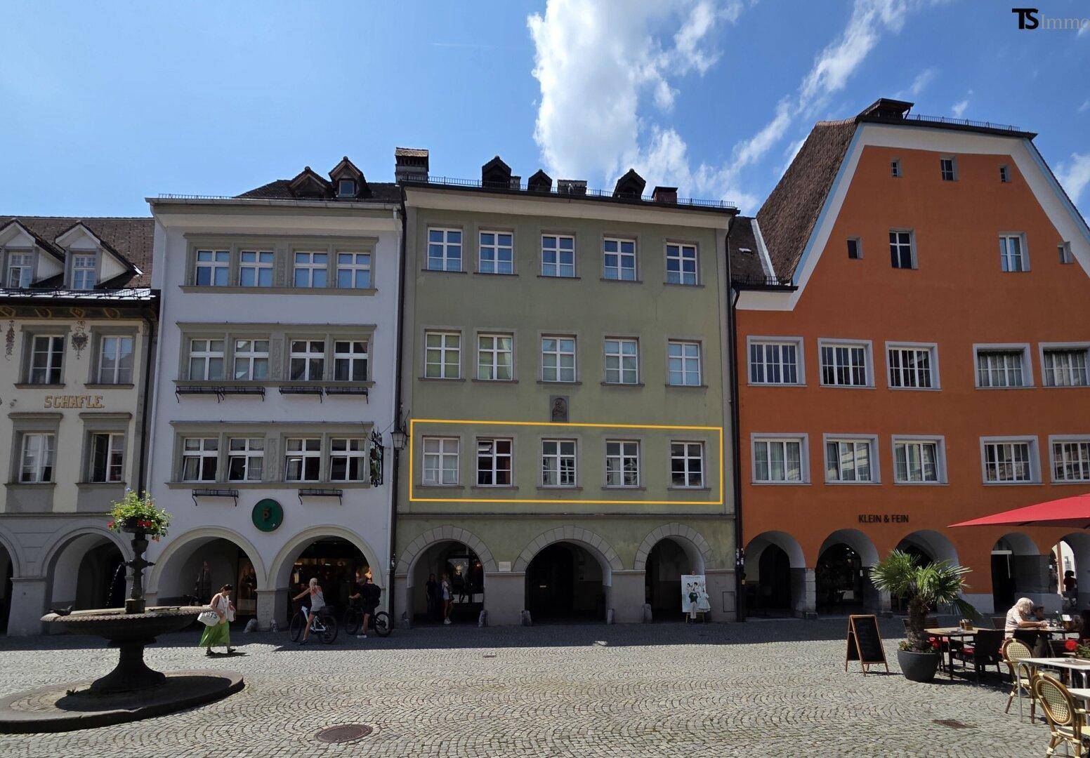 Historisches Gebäude in einer belebten Altstadtgasse mit Geschäften und Cafés, unter blauem Himmel.