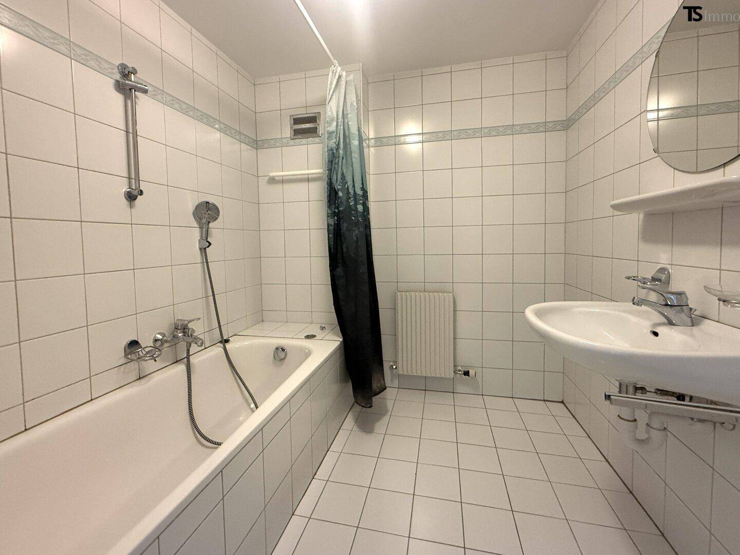 Badezimmer mit Badewanne, Duschvorhang, Waschbecken und Heizkörper, komplett gefliest.