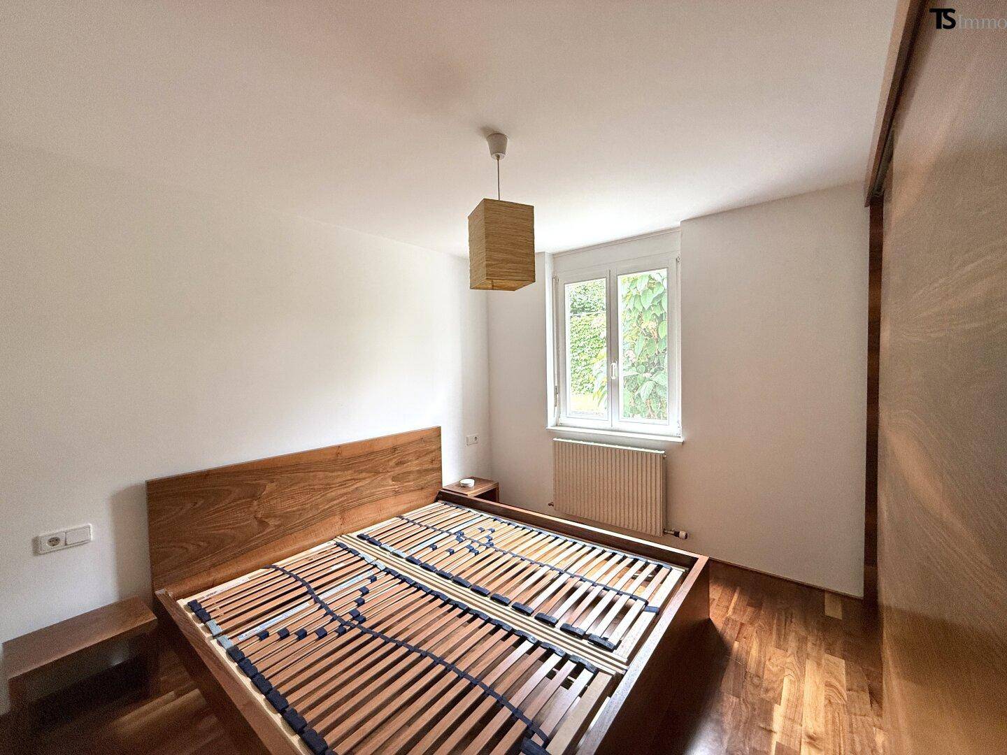 Schlafzimmer mit Holzbettrahmen, Parkettboden und Fenster mit Blick ins Grüne.