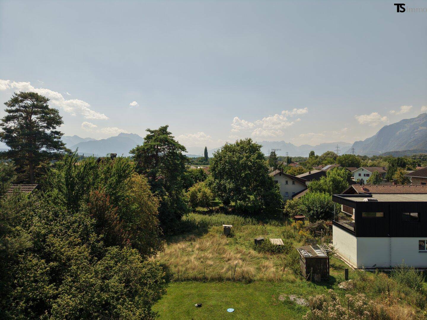 Aussicht auf eine grüne Fläche mit einem modernen Gebäude und Bergen am Horizont unter blauem Himmel.