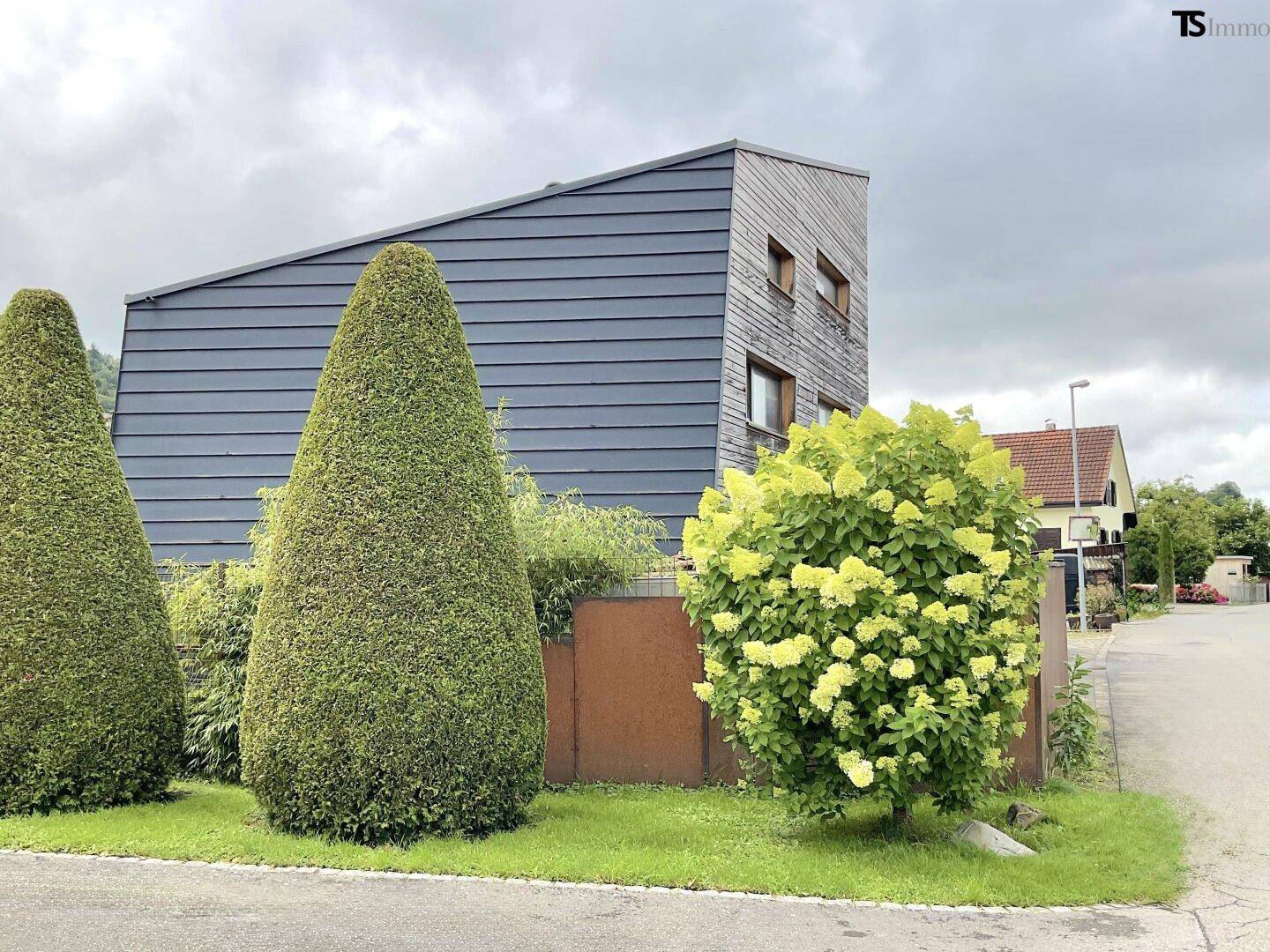 Seitenansicht des modernen Hauses mit Holzfassade und gepflegter Bepflanzung an der Straße.