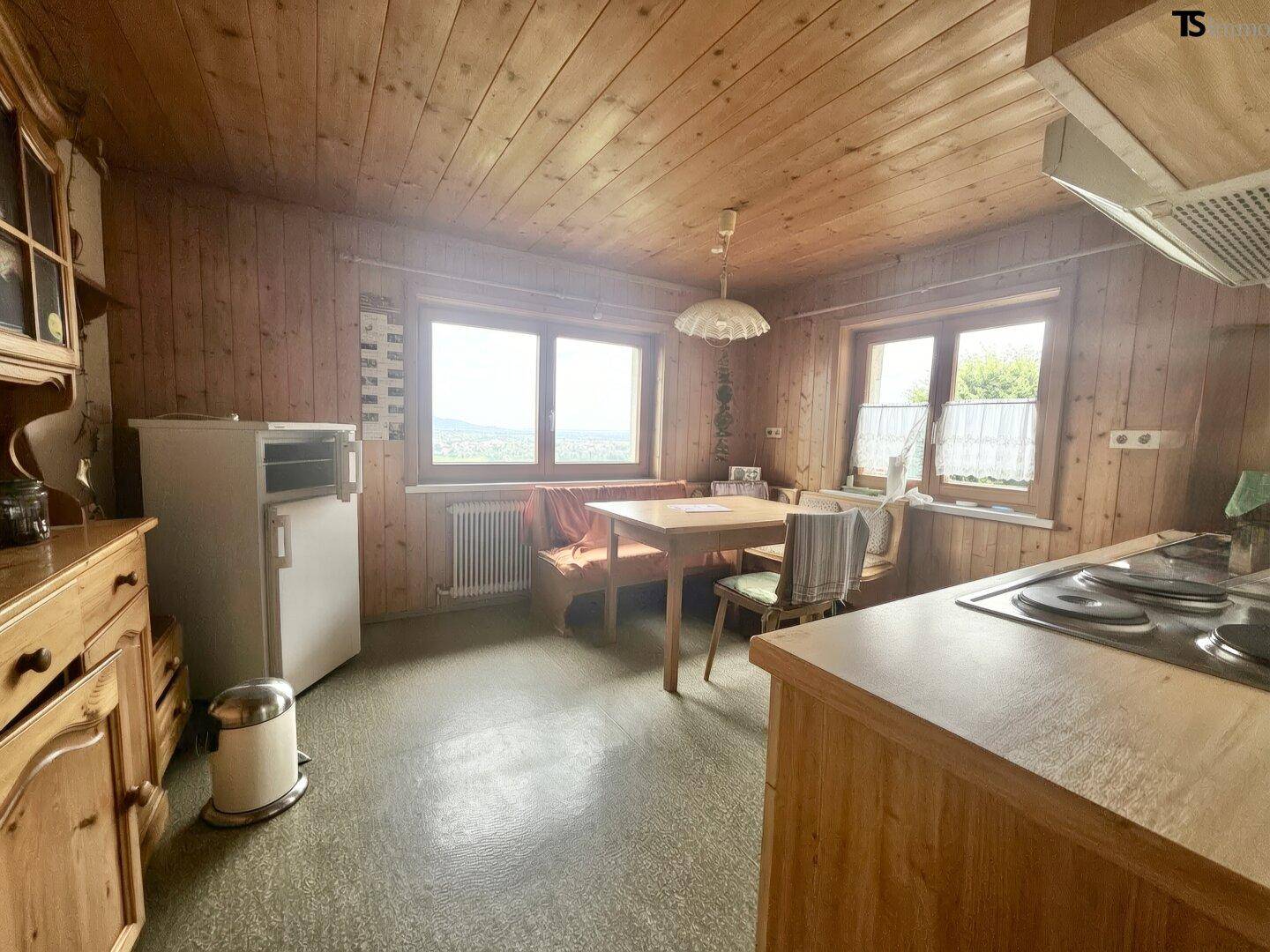 Rustikale Wohnküche mit Holzvertäfelung, Essbereich und Blick ins Grüne durch die Fenster.