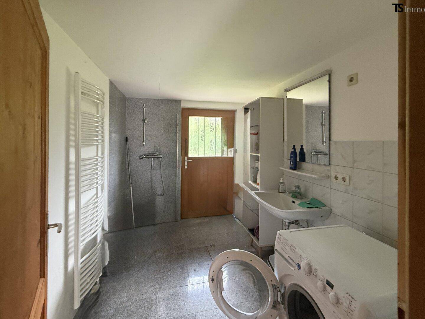Waschküche mit Dusche, Waschmaschine und Fenster zum Außenbereich, gefliestem Boden.