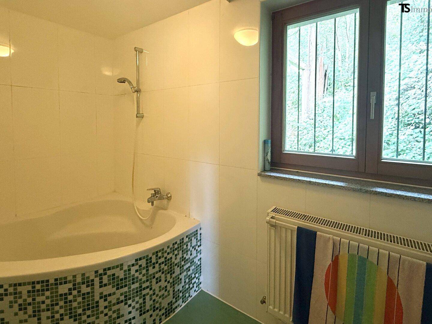 Badezimmer mit Badewanne, Duschvorrichtung und Fenster mit Blick ins Grüne.