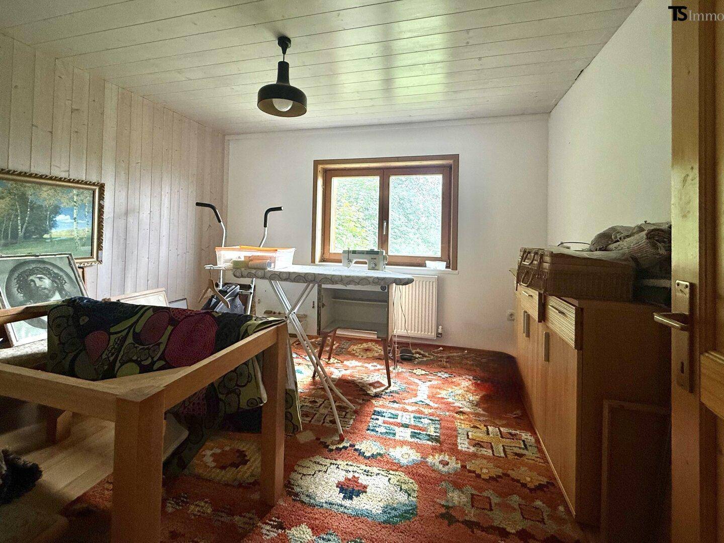 Schlafzimmer mit Teppichboden, Holzvertäfelung und Fenster zum Garten, eingerichtet mit einem Bügelbrett.