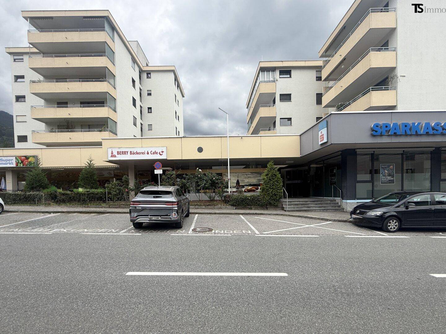 Außenansicht eines Geschäftsgebäudes mit Wohnungen darüber und einem Parkplatz davor.