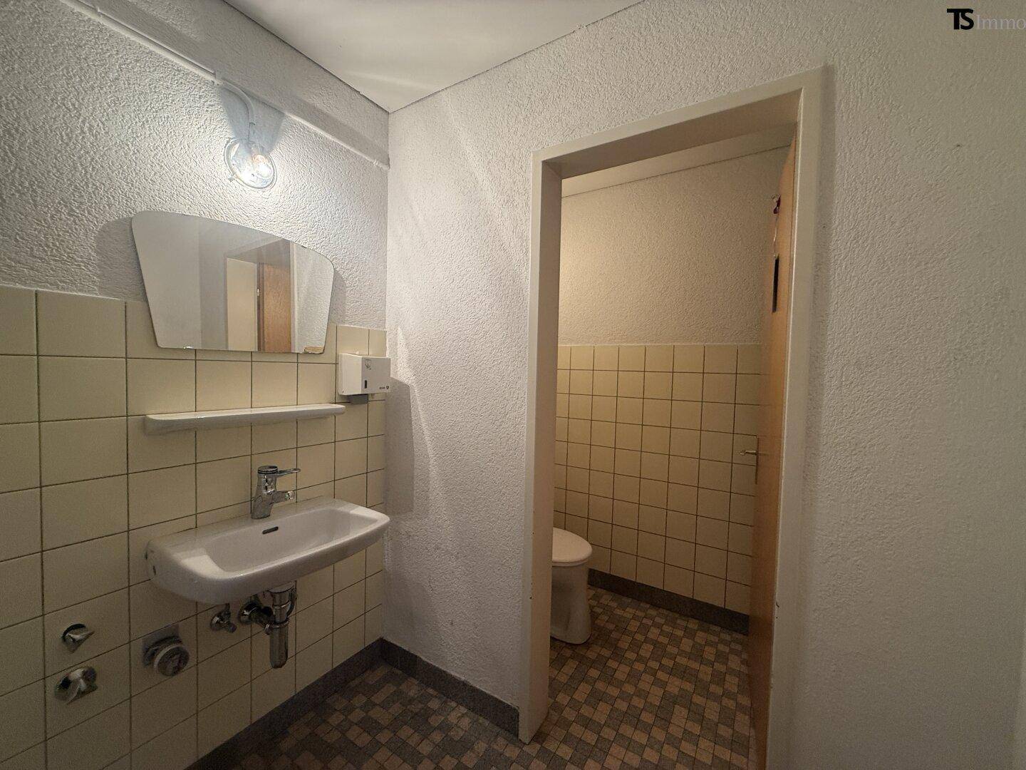 Kleines Badezimmer mit Waschbecken, Spiegel und separater Toilette, ausgestattet mit hellen Fliesen.