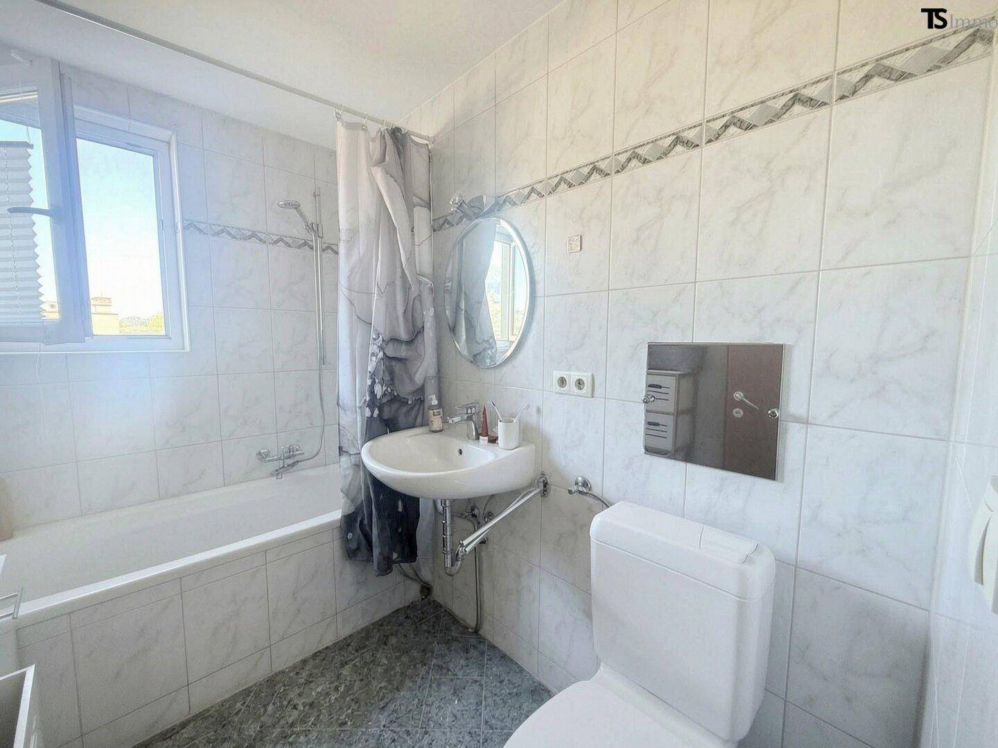 Badezimmer mit weißen Fliesen, Badewanne mit Duschvorhang, Waschbecken und WC, mit Fenster.