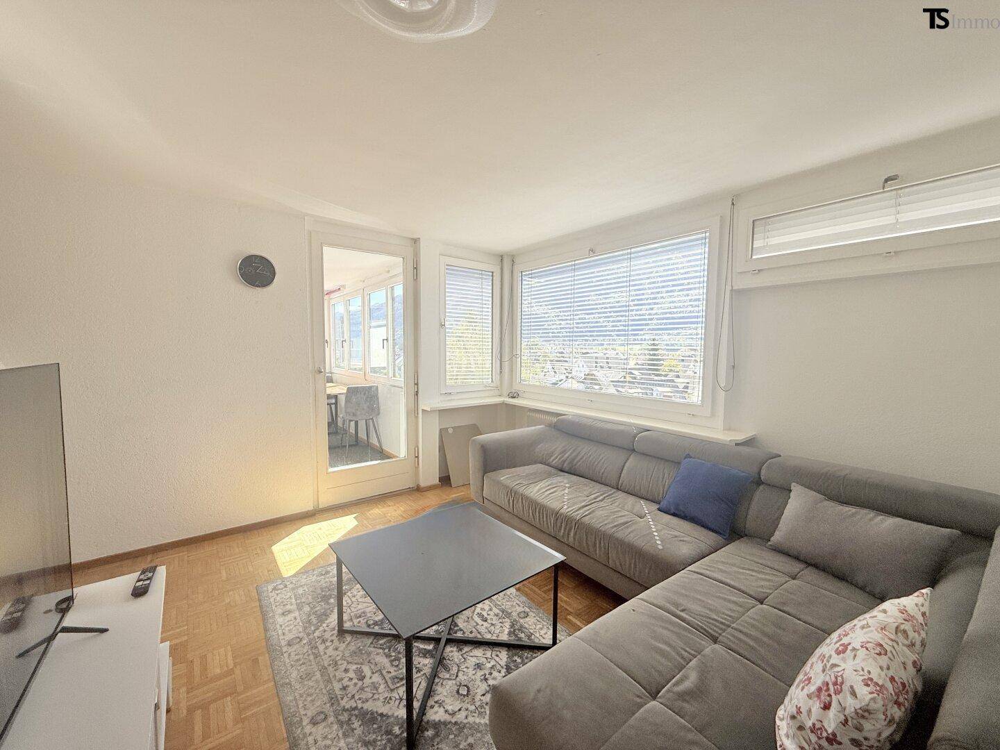 Helles Wohnzimmer mit Sofa, Fernseher und Zugang zu einem weiteren Raum, mit Parkettboden.