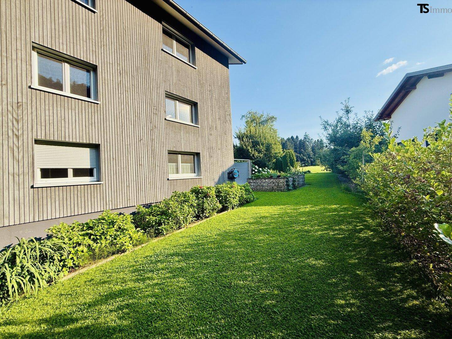 Weitläufiger grüner Rasenbereich entlang der Hausfassade mit Sträuchern und Bäumen im Hintergrund.