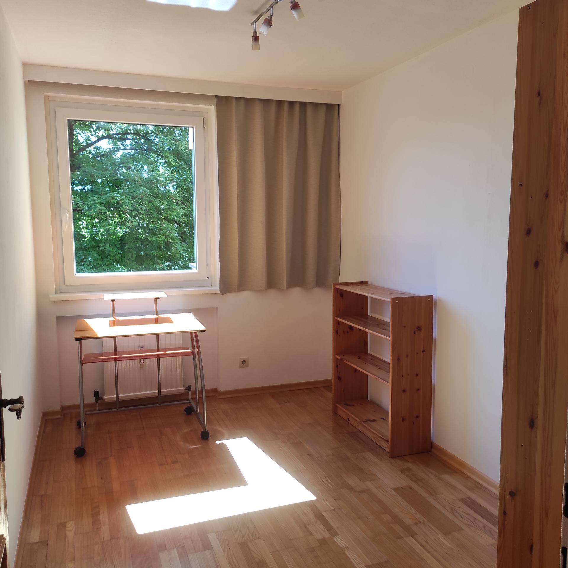 Helles Zimmer mit Holzboden, einem Fenster mit Blick ins Grüne und einem kleinen Arbeitstisch.