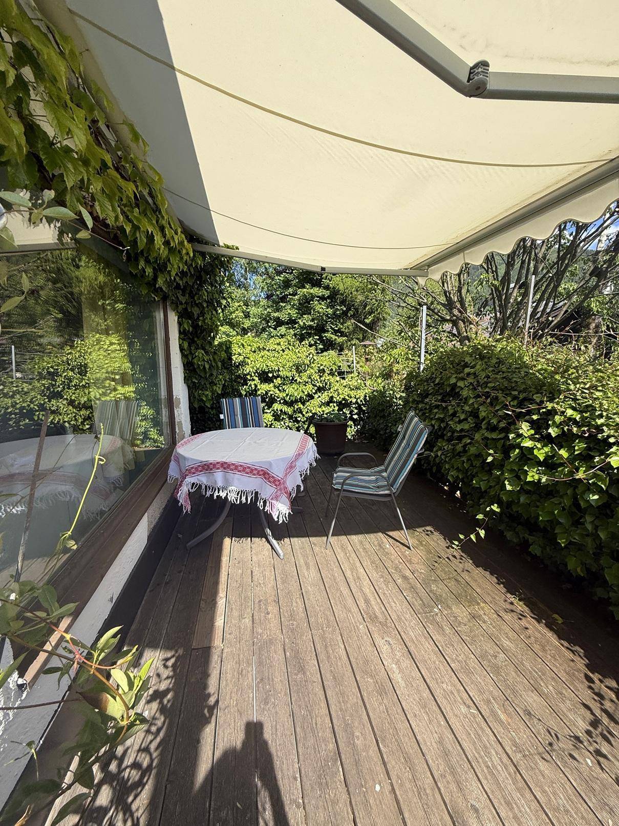 Überdachte Holzterrasse mit Gartenmöbeln und üppiger grüner Bepflanzung.
