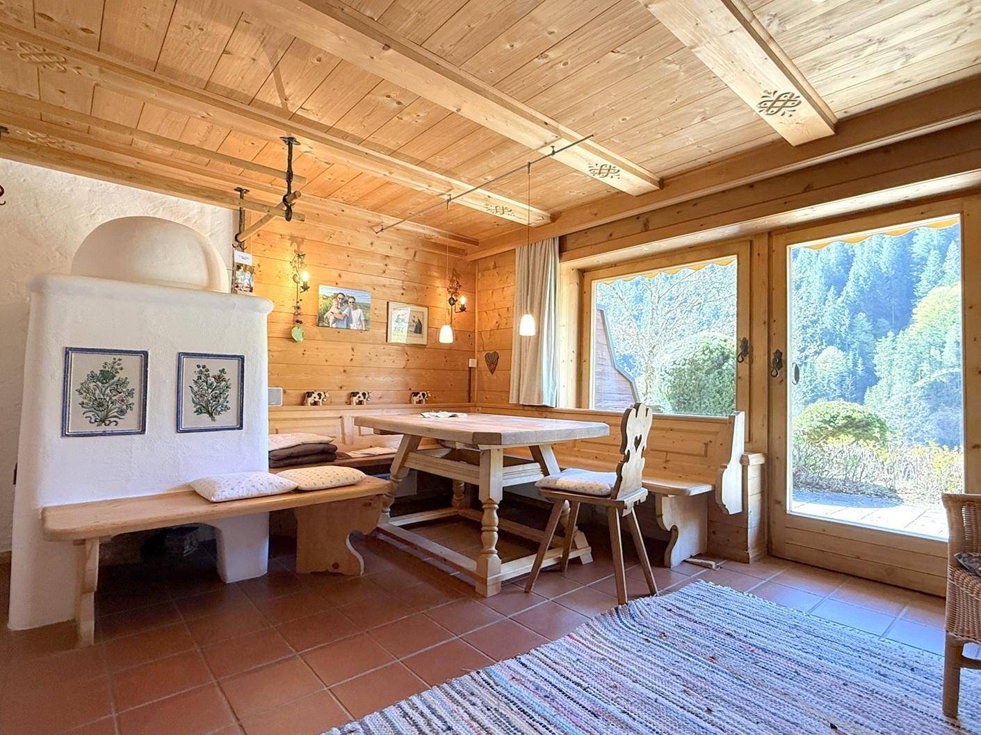 Heller Essbereich mit Holztisch, Eckbank und Kachelofen, umgeben von Holzwänden und großen Fenstern.
