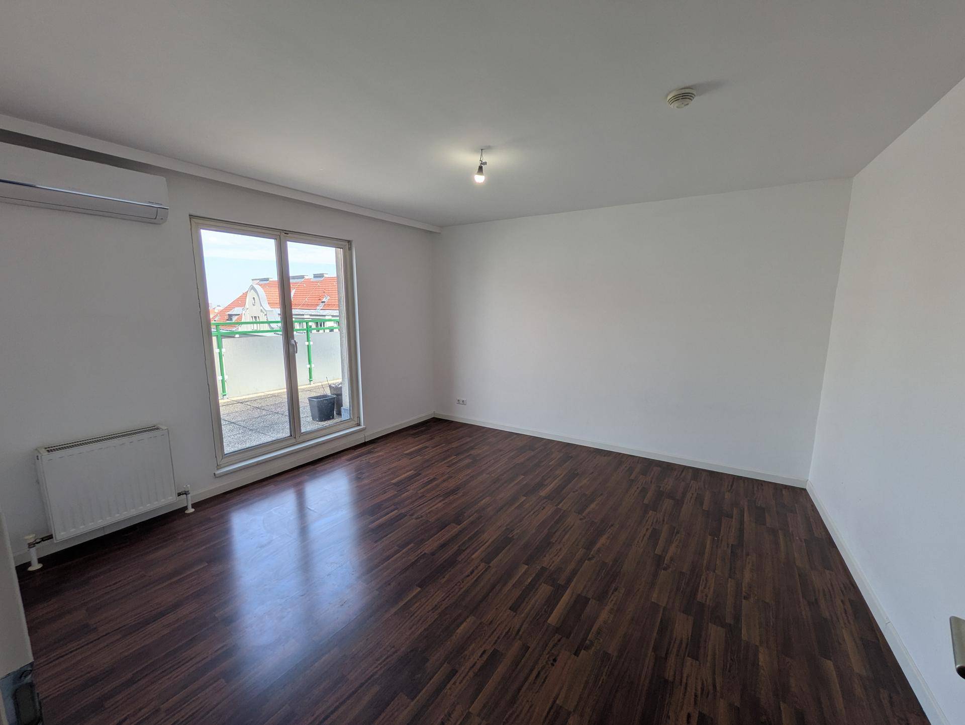 Helles Zimmer mit dunklem Holzboden, Klimaanlage und direktem Zugang zu einer sonnigen Terrasse.