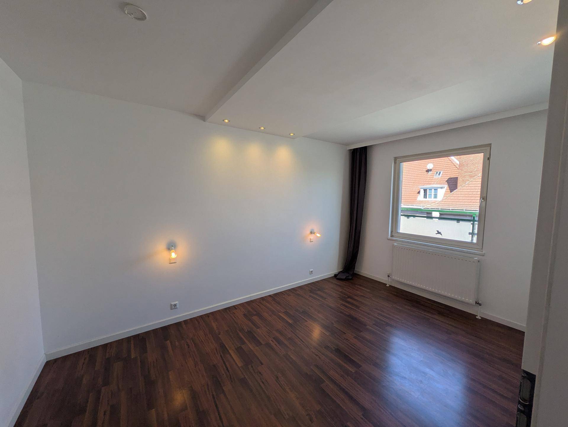 Helles Schlafzimmer mit dunklem Holzboden, weißen Wänden und einem Fenster mit Blick auf die Dächer.