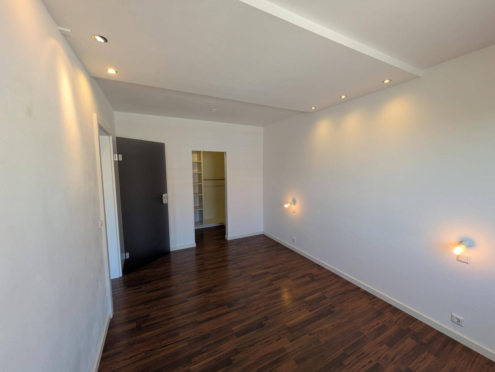 Geräumiges Schlafzimmer mit dunklem Holzboden, weißen Wänden und direktem Zugang zu einem begehbaren Kleiderschrank.