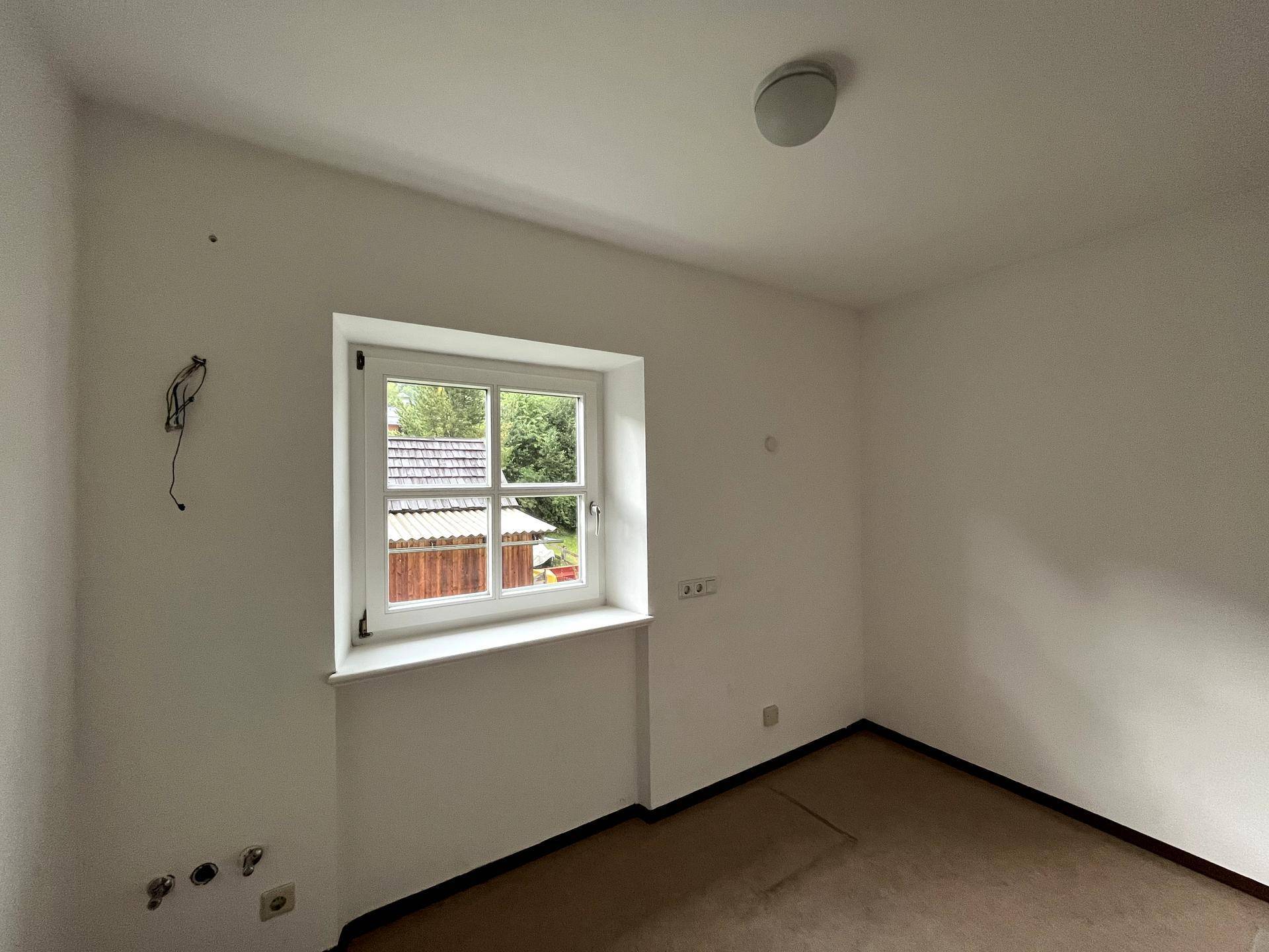 Helles Schlafzimmer mit weiß gestrichenen Wänden und Blick ins Grüne durch das Fenster.