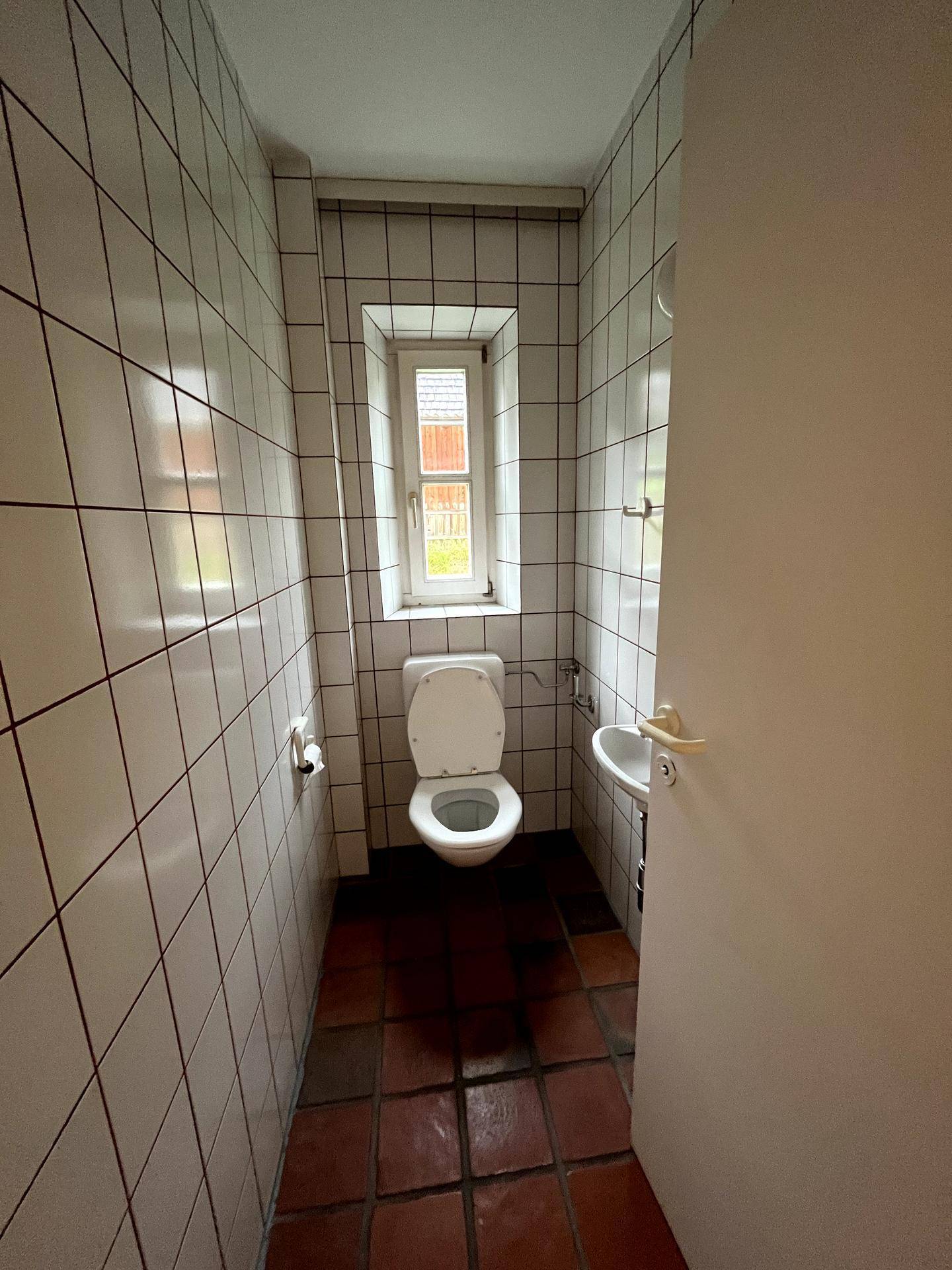 Kleines Gäste-WC mit weißen Fliesen, Terrakottaboden und einem schmalen Fenster.