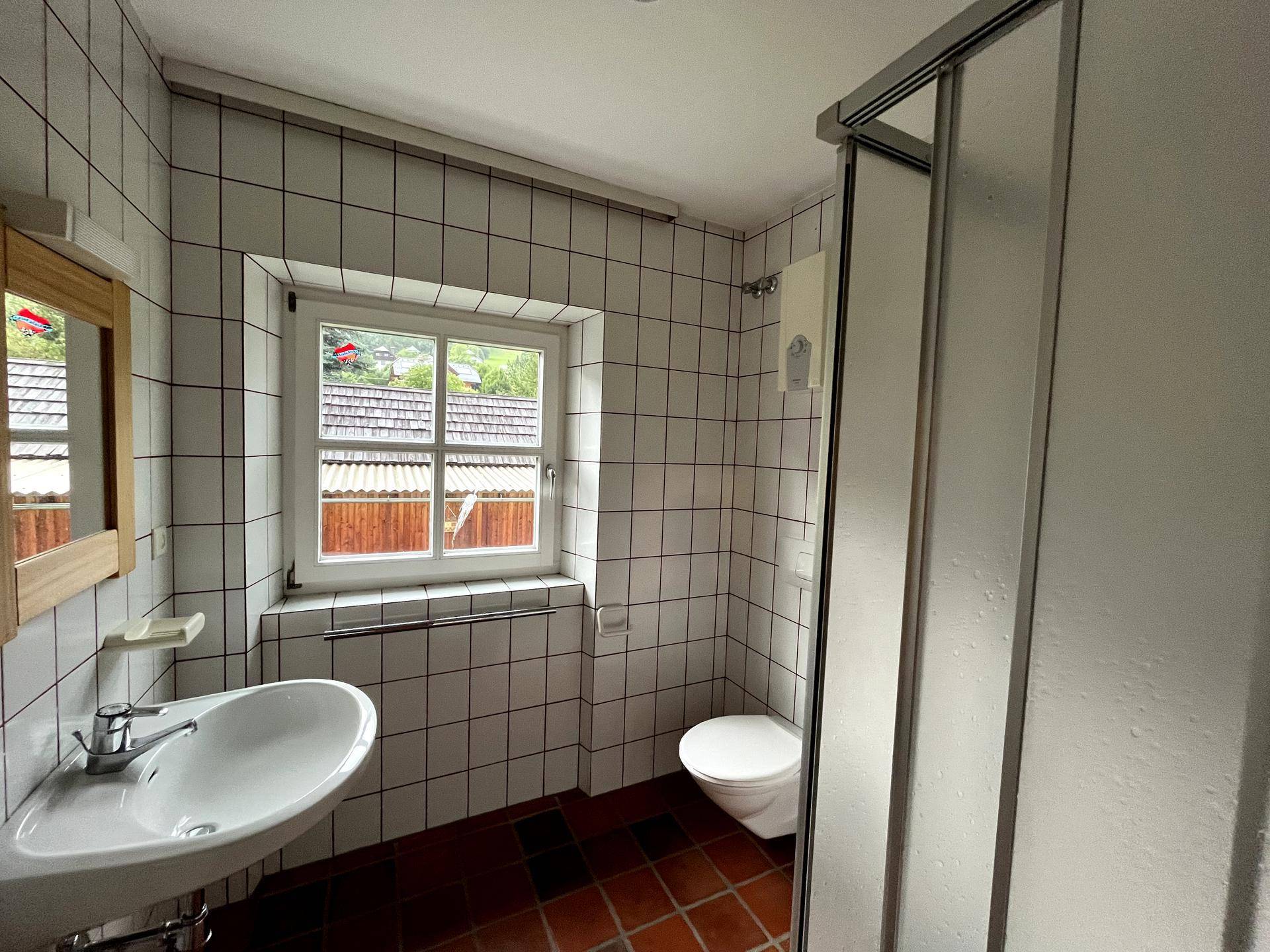Funktionales Badezimmer mit Duschkabine, WC, Waschbecken und Fenster, komplett gefliest.