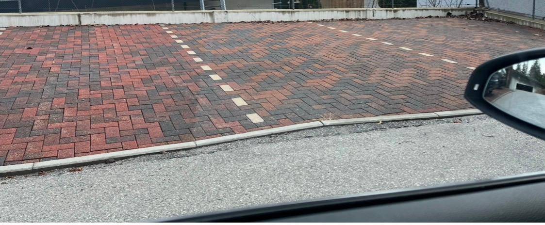 Gepflasterter Außenparkplatz mit roten und grauen Ziegelsteinen und weißen Stellplatzmarkierungen, teilweise von einem Auto verdeckt.