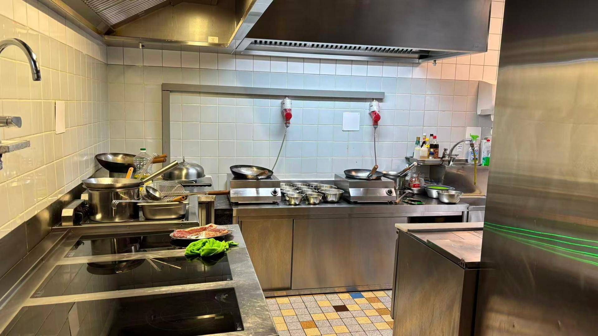 Professionelle Küche mit Edelstahlgeräten, Induktionskochfeld und gefliesten Wänden.