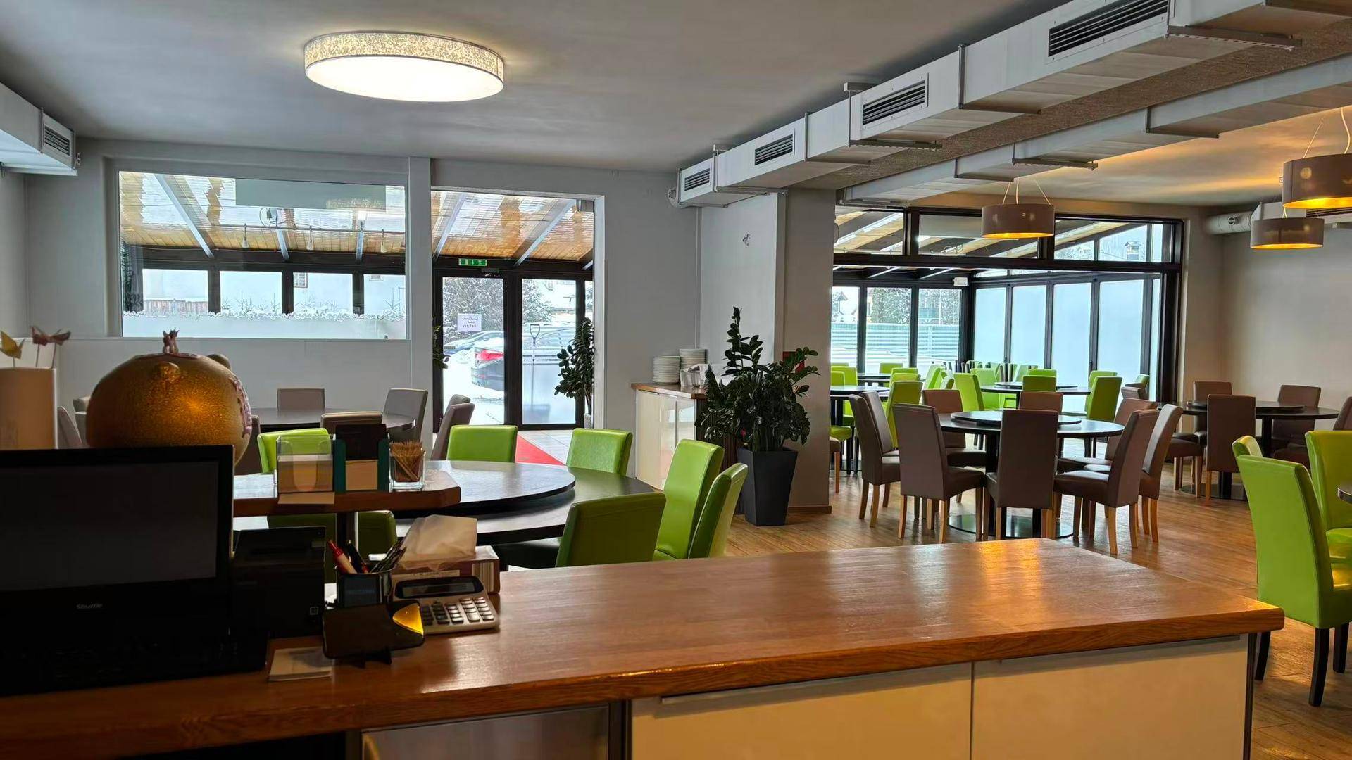 Heller Restaurantbereich mit runden Tischen, grünen Stühlen und großen Fenstern zur Terrasse.