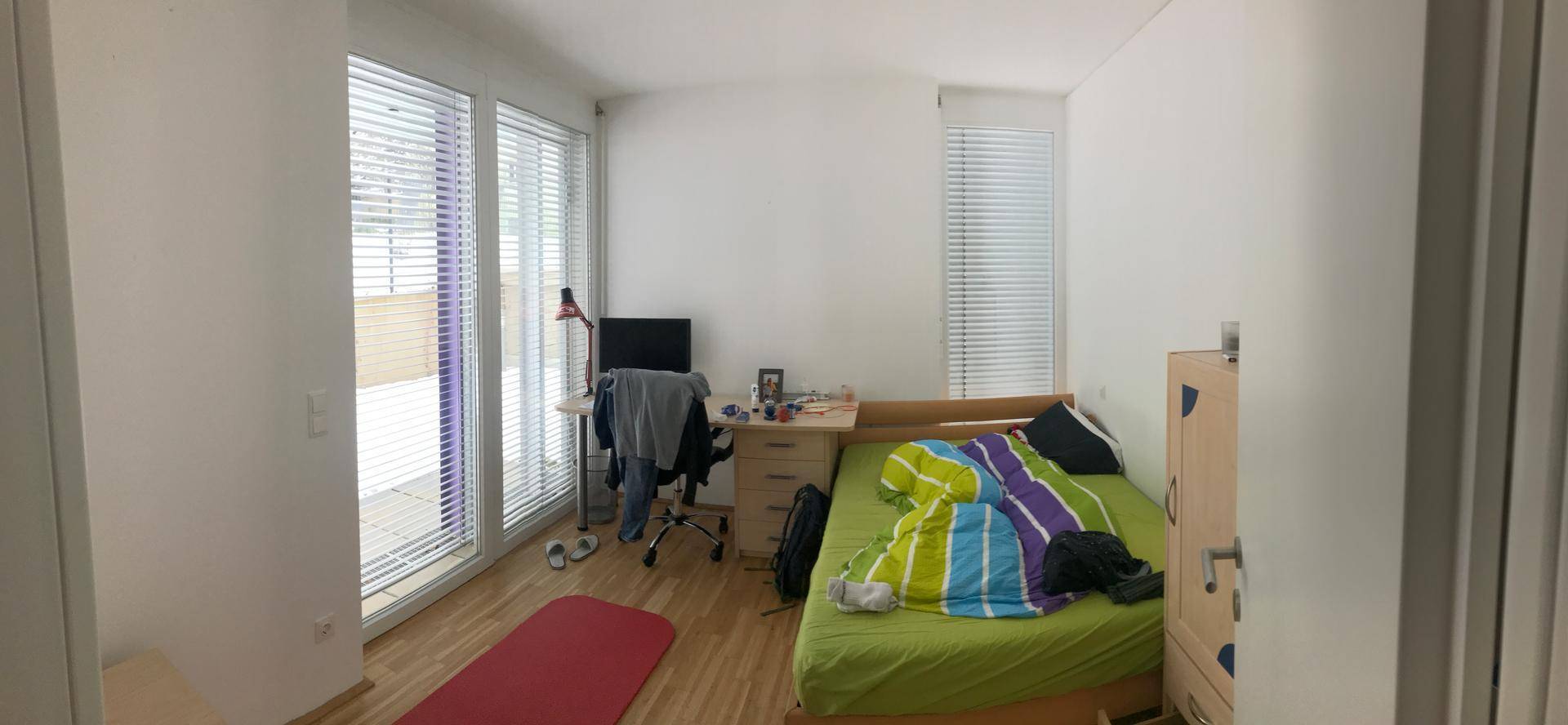 Helles Schlafzimmer mit Holzboden, Schreibtisch und großen Fenstern mit Jalousien.
