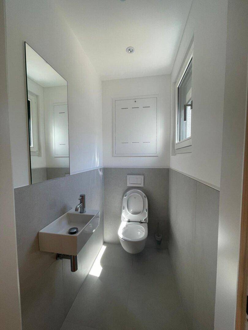 Modernes Gäste-WC mit Wand-WC, kleinem Waschbecken und Fenster für natürliche Belüftung.