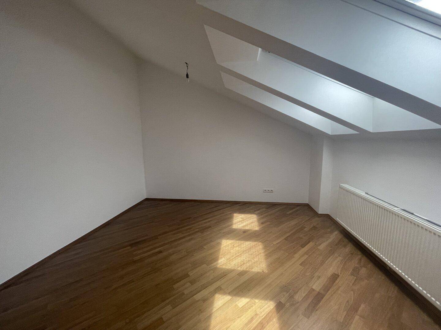 Helles Zimmer im Dachgeschoss mit Holzboden, Dachfenstern und Heizkörper.