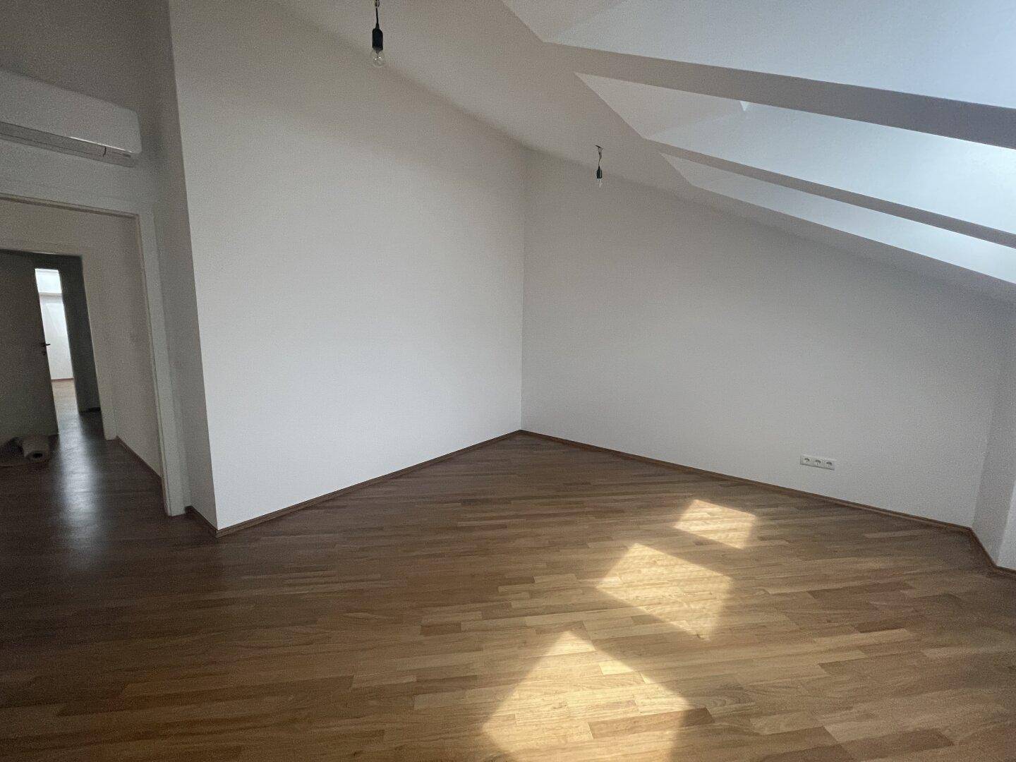 Leeres Zimmer im Dachgeschoss mit Holzboden, schrägen Wänden und Klimaanlage.