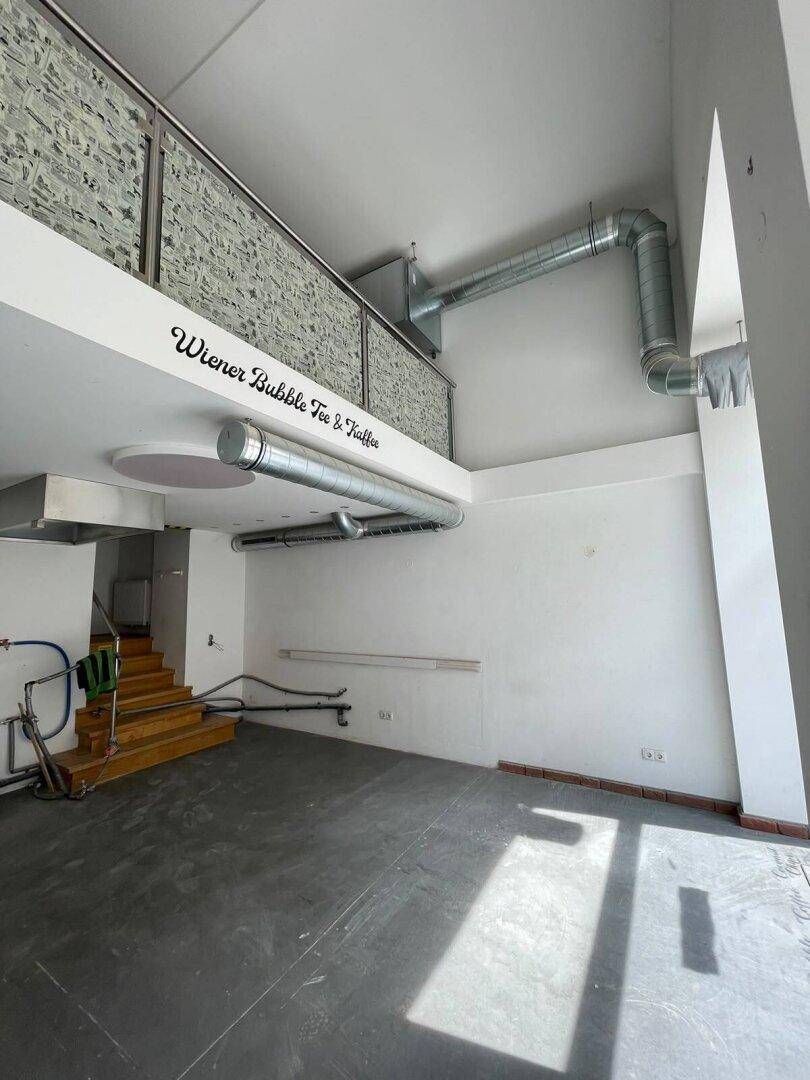 Innenansicht eines Ladenlokals mit hoher Decke, sichtbaren Lüftungsrohren und einer Galerie im Hintergrund.